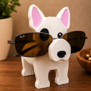 May include: A white dog-shaped eyeglass holder with pink inner ears and a black nose. The holder is displaying a pair of Ray-Ban sunglasses with brown lenses. The holder is made of a smooth, glossy material and sits on a wooden surface.
