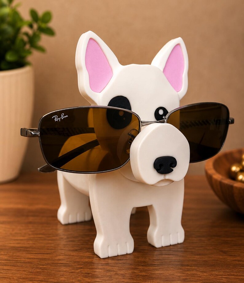 May include: A white dog-shaped eyeglass holder with pink inner ears and a black nose. The holder is displaying a pair of Ray-Ban sunglasses with brown lenses. The holder is made of a smooth, glossy material and sits on a wooden surface.