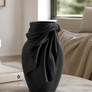 May include: A matte black ceramic vase with a draped fabric design around the neck. The vase has a rounded body and a smooth, elegant finish. Home decor item.