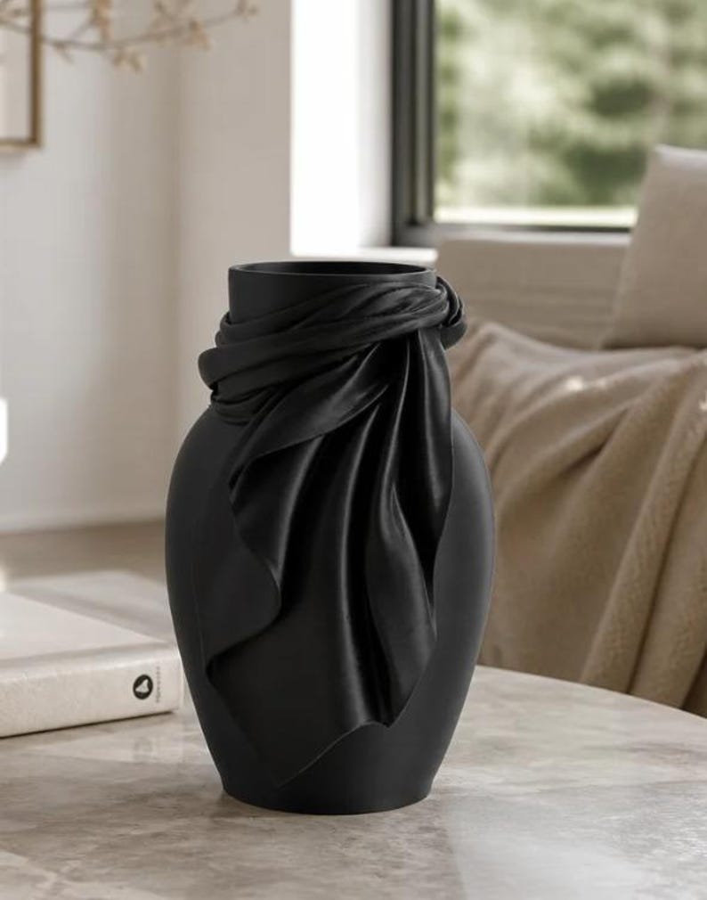 May include: A matte black ceramic vase with a draped fabric design around the neck. The vase has a rounded body and a smooth, elegant finish. Home decor item.
