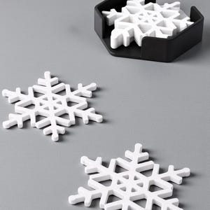 Puede incluir: Posavasos blancos con forma de copo de nieve con un soporte negro. Dos posavasos est&aacute;n colocados sobre una superficie gris, con un juego de posavasos en un soporte negro. Tema de vacaciones de invierno.