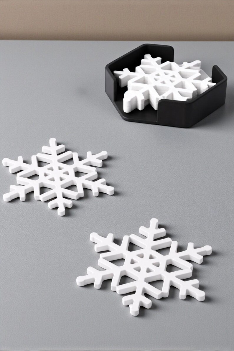 Puede incluir: Posavasos blancos con forma de copo de nieve con un soporte negro. Dos posavasos est&aacute;n colocados sobre una superficie gris, con un juego de posavasos en un soporte negro. Tema de vacaciones de invierno.