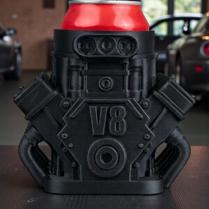 May include: A black V8 engine-shaped can cooler holds a red soda can. The cooler has detailed mechanical features and the text "V8" on the front. The design is a unique novelty item.