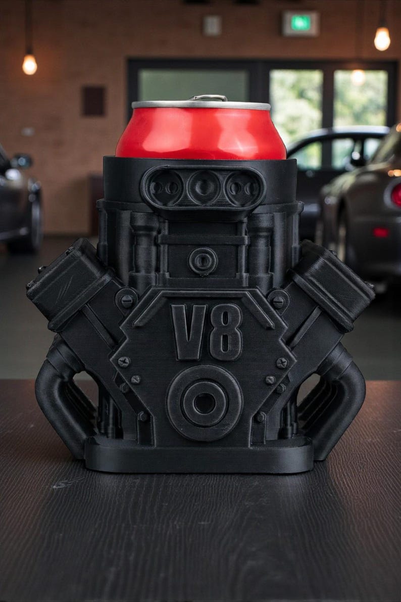 May include: A black V8 engine-shaped can cooler holds a red soda can. The cooler has detailed mechanical features and the text "V8" on the front. The design is a unique novelty item.