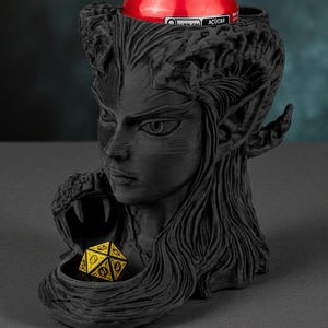 May include: A black, 3D-printed fantasy-themed drink holder featuring a woman's face with long hair and pointed ears. A red can sits in the holder's top, and a yellow and black die rests in a lower compartment. The holder is designed for tabletop gaming.