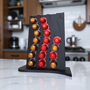 May include: A black coffee pod holder with a curved design, holding various coffee pods in gold and red. The holder has a coffee cup icon and is displayed on a white countertop.