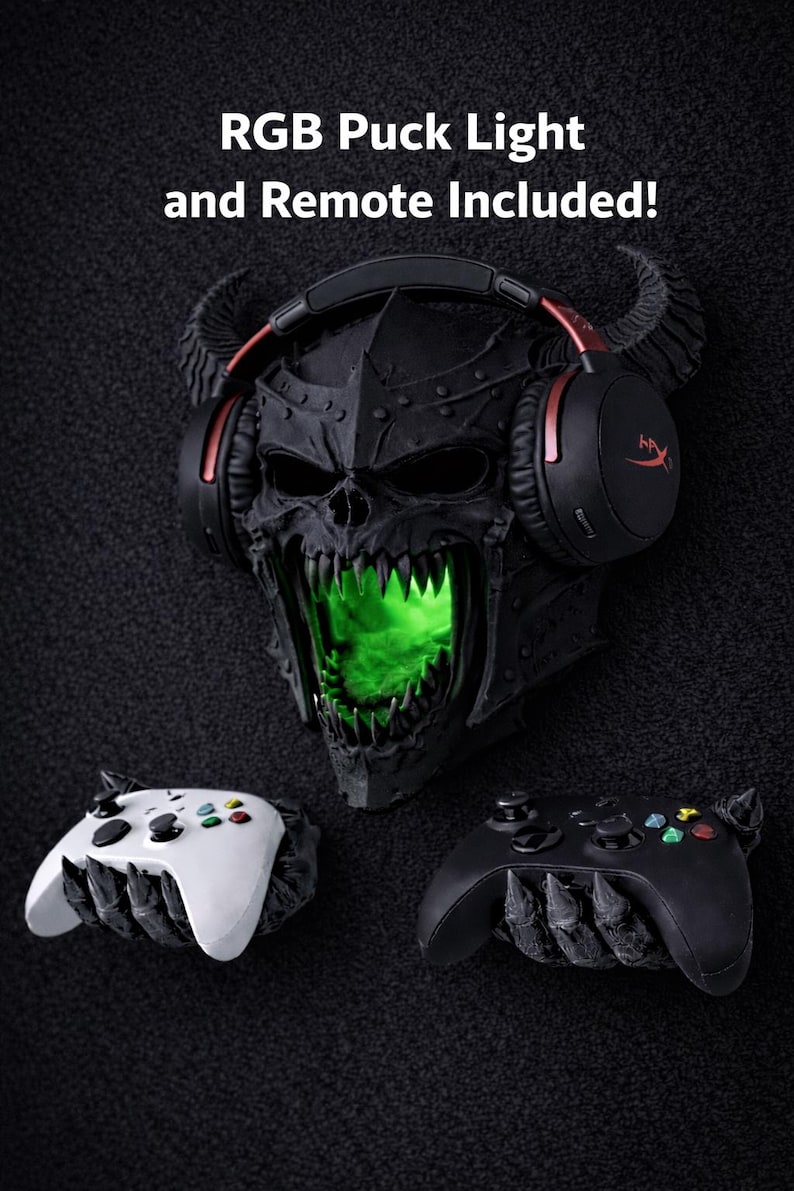 May include: A black skull-shaped wall mount with a green glowing mouth, holding a pair of black and red gaming headphones. Two game controllers, one white and one black, are positioned below. The text "RGB Puck Light and Remote Included!" is displayed above.
