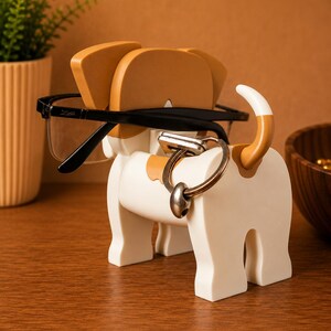May include: A white and tan dog-shaped eyeglasses and key holder. The holder has a black pair of eyeglasses resting on the dog's head and a silver key ring attached to the dog's body. The dog has tan ears and a tan and white tail.