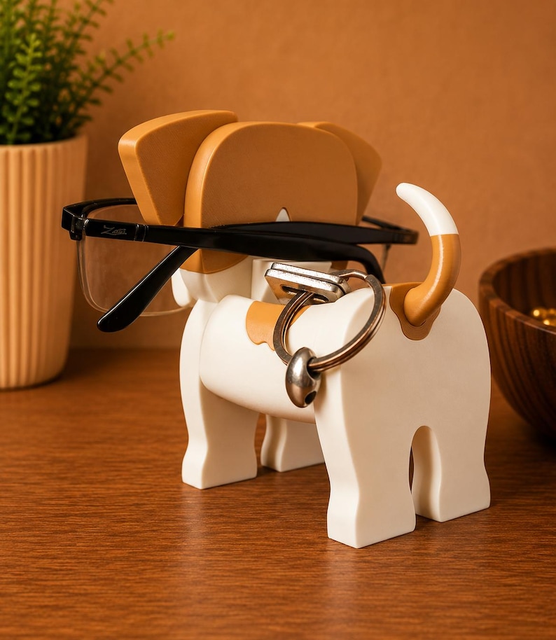 May include: A white and tan dog-shaped eyeglasses and key holder. The holder has a black pair of eyeglasses resting on the dog's head and a silver key ring attached to the dog's body. The dog has tan ears and a tan and white tail.