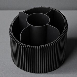 May include: A black desk organizer with a cylindrical shape and vertical ribbed exterior. The interior is divided into multiple compartments, including a central circular section. The organizer is made of a matte material.
