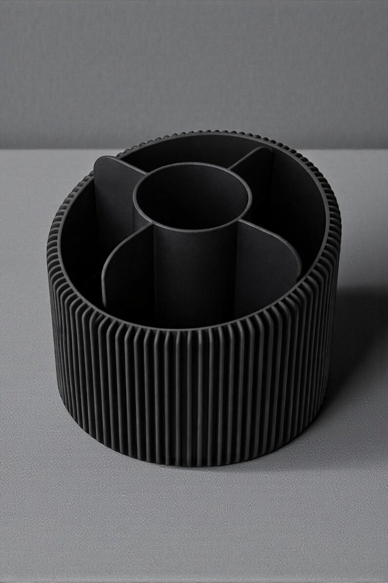 May include: A black desk organizer with a cylindrical shape and vertical ribbed exterior. The interior is divided into multiple compartments, including a central circular section. The organizer is made of a matte material.