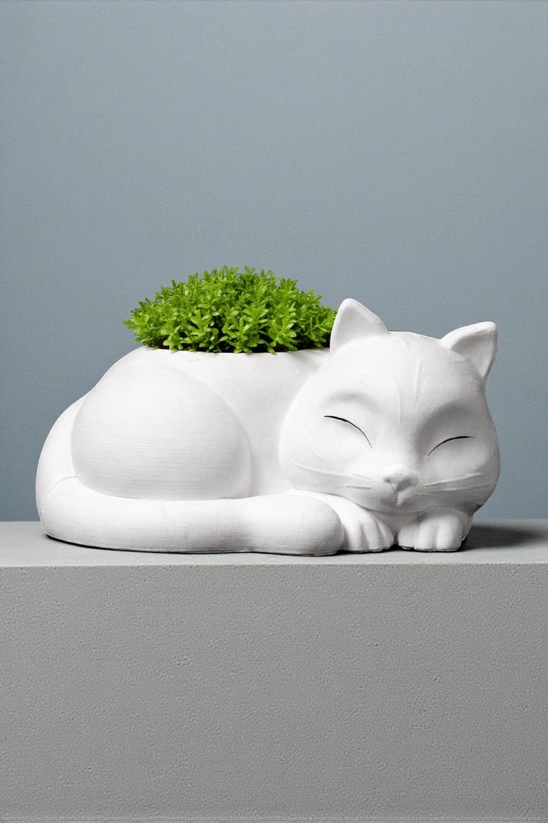May include: A white ceramic cat-shaped planter with a sleeping pose. The planter is filled with green plants. The cat's eyes are closed, and it rests on a gray surface against a blue background. A decorative item for home decor.