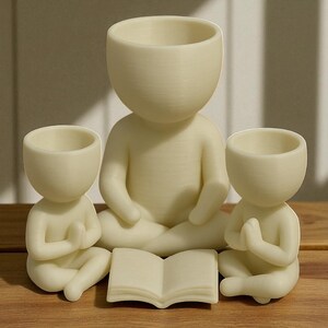 May include: Three cream-colored, human-shaped planters in a meditative pose. The largest planter sits in the center, with two smaller planters on either side. A small, open book rests in front of the central figure. The planters are on a wooden surface.
