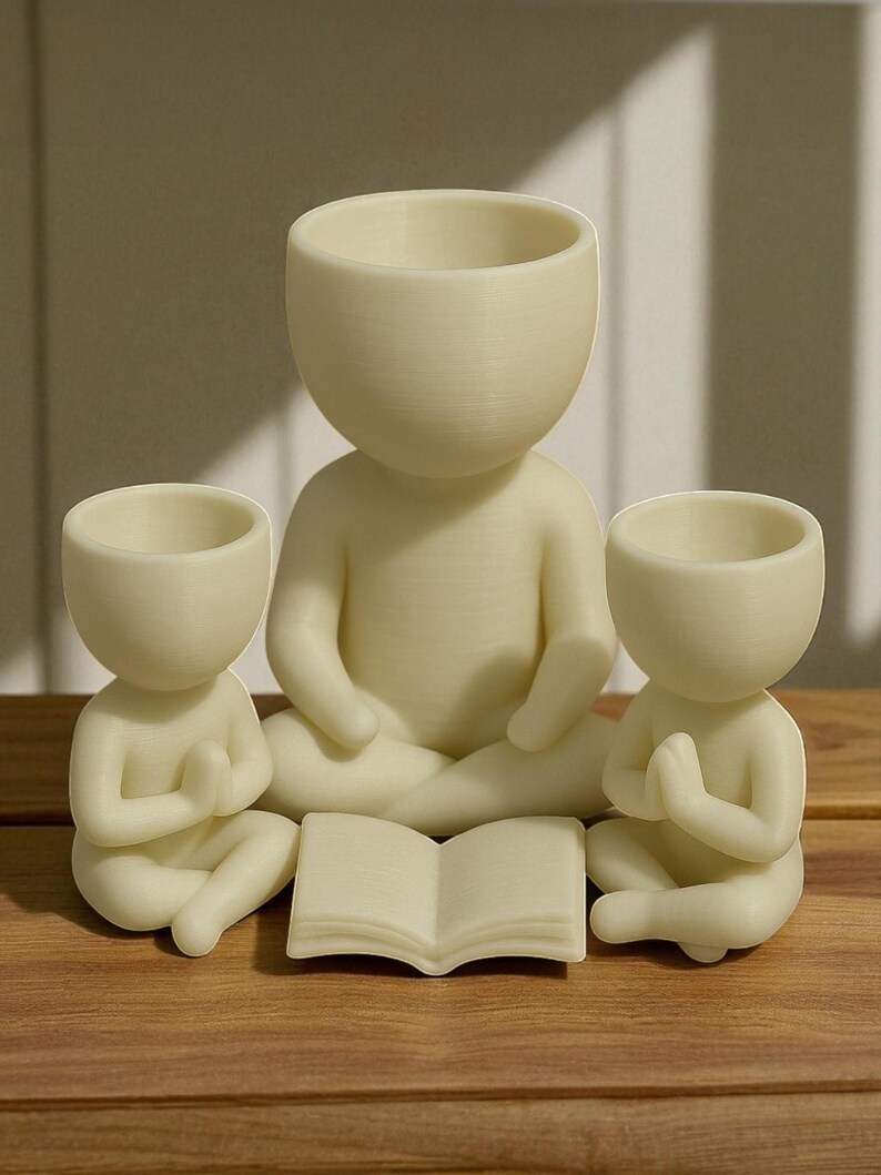 May include: Three cream-colored, human-shaped planters in a meditative pose. The largest planter sits in the center, with two smaller planters on either side. A small, open book rests in front of the central figure. The planters are on a wooden surface.