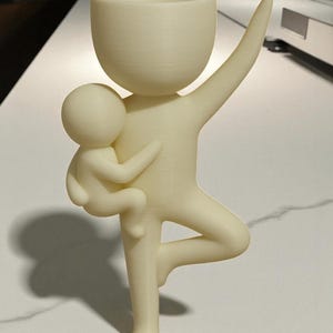 May include: A cream-colored, stylized figurine of a parent figure holding a child. The parent figure has one arm raised and is balancing on one leg. The top of the parent figure is a cup-shaped planter. The child is cradled in the parent's other arm.