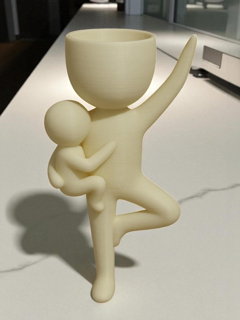 May include: A cream-colored, stylized figurine of a parent figure holding a child. The parent figure has one arm raised and is balancing on one leg. The top of the parent figure is a cup-shaped planter. The child is cradled in the parent's other arm.