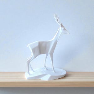 May include: A white, geometric deer sculpture with detailed antlers stands on a white base. The minimalist design features sharp angles and clean lines, creating a modern aesthetic. The sculpture is displayed on a light wood shelf against a pale blue wall.
