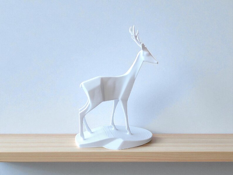 May include: A white, geometric deer sculpture with detailed antlers stands on a white base. The minimalist design features sharp angles and clean lines, creating a modern aesthetic. The sculpture is displayed on a light wood shelf against a pale blue wall.