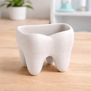 May include: A white, tooth-shaped planter with three rounded roots. The planter has a rectangular opening at the top and is made of a textured material. The planter is sitting on a light-coloured wooden surface.