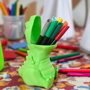 K&ouml;nnte beinhalten: Ein hellgr&uuml;ner, dinosaurierf&ouml;rmiger Stifthalter, gef&uuml;llt mit bunten Markern. Der Stifthalter hat eine breite &Ouml;ffnung und ein strukturiertes Design. Weitere Stifte und Marker sind im Hintergrund verstreut.