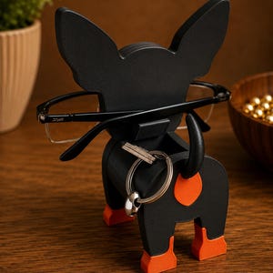 May include: A black dog-shaped eyeglasses holder with orange accents on the ears, paws, and rear. The holder is designed to hold eyeglasses and a set of keys. The eyeglasses are black-framed. The holder is made of wood.