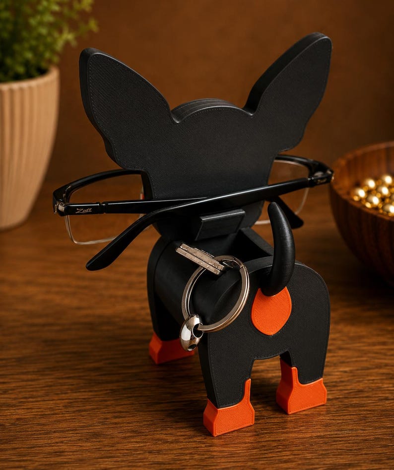 May include: A black dog-shaped eyeglasses holder with orange accents on the ears, paws, and rear. The holder is designed to hold eyeglasses and a set of keys. The eyeglasses are black-framed. The holder is made of wood.