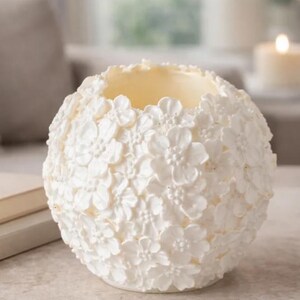May include: A white, spherical candle holder with a floral design. The surface is covered in small, three-dimensional flowers. The interior is a light yellow color, and the holder is sitting on a neutral surface. The candle holder is a decorative home accent.