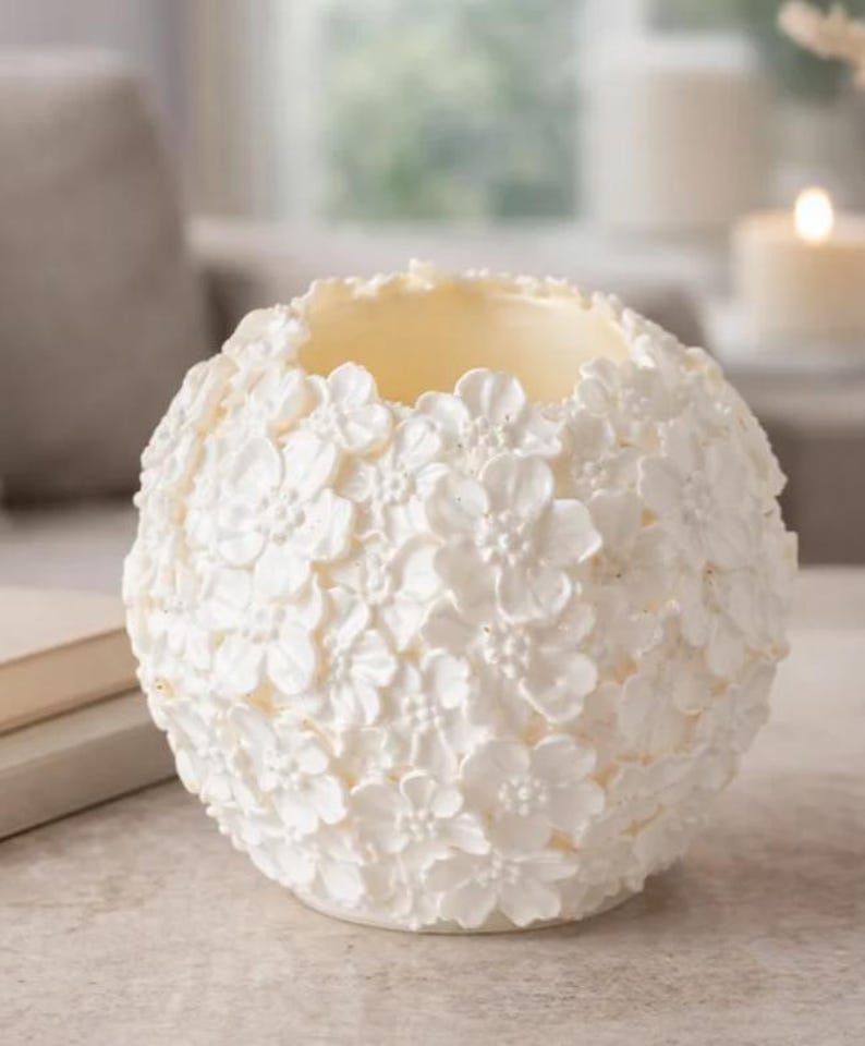 May include: A white, spherical candle holder with a floral design. The surface is covered in small, three-dimensional flowers. The interior is a light yellow color, and the holder is sitting on a neutral surface. The candle holder is a decorative home accent.
