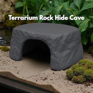 May include: A gray, rock-shaped terrarium hide cave. The cave has a large arched opening and a textured surface. The text "Terrarium Rock Hide Cave" is displayed in white. The cave is surrounded by greenery and sand.
