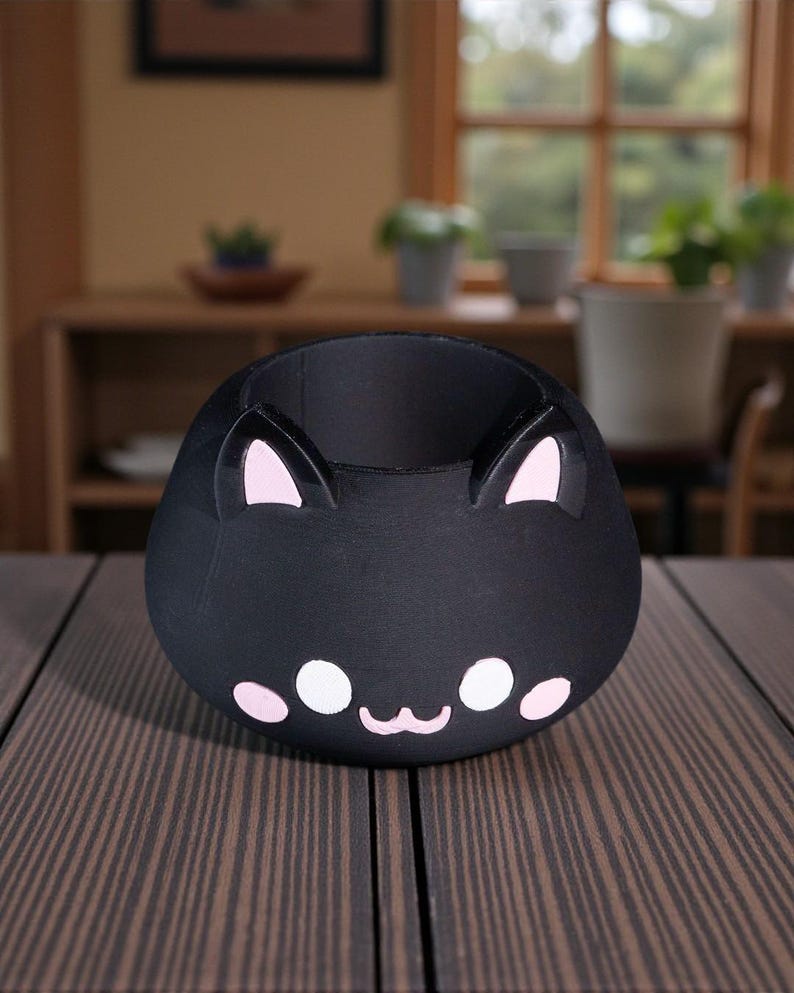 K&ouml;nnte beinhalten: Ein schwarzer, katzenf&ouml;rmiger Beh&auml;lter mit rosa Akzenten. Der Beh&auml;lter hat einen runden K&ouml;rper mit zwei aufrechten Ohren und einem Gesichtsdesign mit wei&szlig;en und rosa Details. Der Beh&auml;lter steht auf einer braunen Holzoberfl&auml;che.