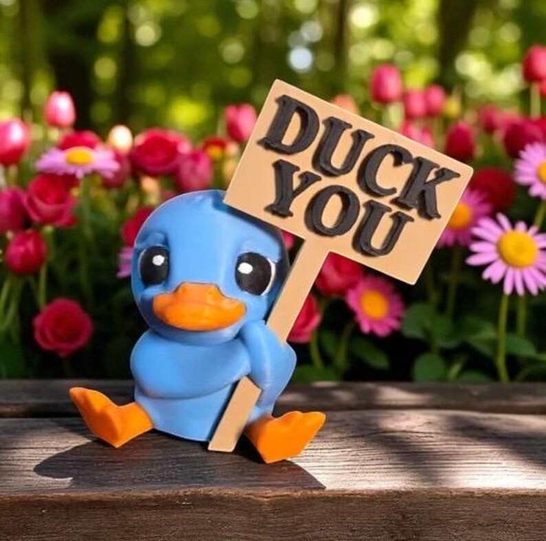 May include: A blue duck figurine with an orange beak and feet, holding a sign that reads "DUCK YOU" in black letters. The duck is sitting on a wooden surface, with a blurred background of flowers.