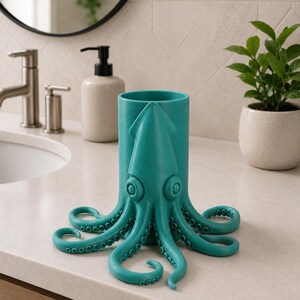 May include: A teal octopus-shaped holder, likely for bathroom accessories, sits on a white countertop. The holder features a cylindrical body with an octopus design, including tentacles and large eyes. A sink, mirror, and plant are in the background.