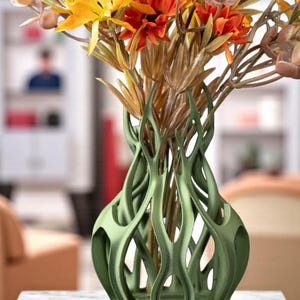May include: A green, open-weave vase containing a bouquet of artificial flowers. The flowers are a mix of orange, yellow, and brown, with varied textures. The vase has a unique, organic design.