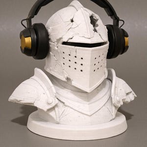 Puede incluir: Un busto blanco de un casco y armadura de caballero, con unos auriculares negros y dorados encima. El casco tiene un aspecto agrietado y desgastado. El busto est&aacute; sobre una base redonda.