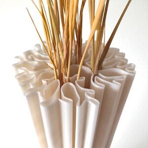 May include: A cream-colored, ruffled vase holds dried, golden-brown grasses. The vase has a unique, wavy design, with vertical, curved sections creating a textured surface. The arrangement is set against a plain white background.
