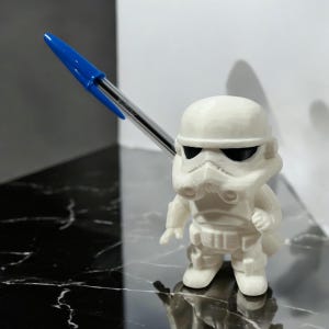 May include: A white Stormtrooper figurine with black eye details, standing upright. A blue and clear pen is positioned above the figurine. The figurine is on a reflective black and white surface.