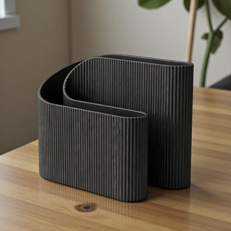 May include: Two dark gray, ribbed vases of different sizes sit on a wooden table. The vases have a modern, minimalist design with vertical lines. One vase curves inward, creating an interesting shape. The background is neutral, with a glimpse of a plant.