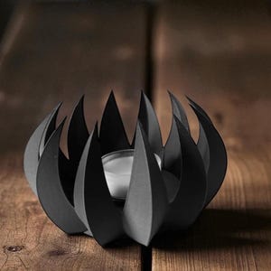 May include: A black metal candle holder with a unique, stylized design. The holder features a series of pointed, flame-like shapes surrounding a central space for a small candle. The candle holder is sitting on a wooden surface.