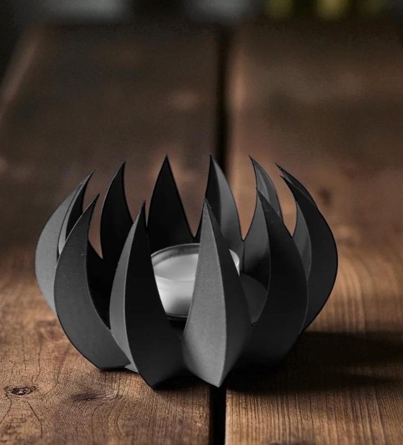 May include: A black metal candle holder with a unique, stylized design. The holder features a series of pointed, flame-like shapes surrounding a central space for a small candle. The candle holder is sitting on a wooden surface.