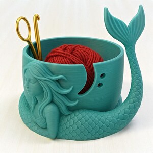 May include: A teal mermaid-themed yarn bowl with a red yarn ball inside. The bowl has a mermaid tail and a sculpted mermaid figure. A gold crochet hook and scissors are placed inside the bowl. The bowl is designed for knitting and crochet enthusiasts.