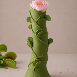 May include: A green vase with a textured surface and vine-like detailing, holding a pink and white lotus flower. The vase has a flared base and is approximately 10 inches tall.