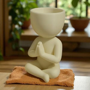 Puede incluir: Una maceta de color crema con forma de persona en posición de meditación sentada. La figura tiene las manos juntas en oración y está sentada sobre una pequeña esterilla naranja texturizada. La cabeza de la figura es un cuenco grande y abierto para plantar.