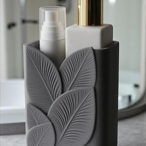 May include: A gray soap dispenser set with a leaf design. The set includes a white pump bottle with a gold dispenser and a white bottle with a clear cap. The set is designed to hold liquid soap and lotion.