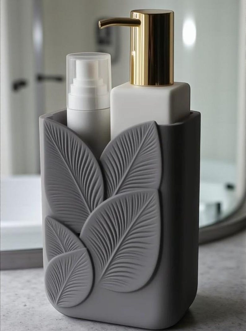 May include: A gray soap dispenser set with a leaf design. The set includes a white pump bottle with a gold dispenser and a white bottle with a clear cap. The set is designed to hold liquid soap and lotion.