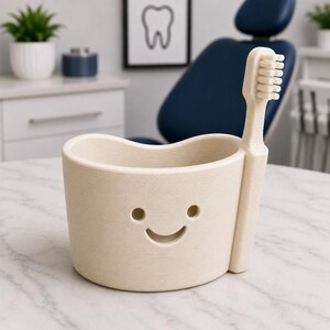 May include: Beige toothbrush holder with a smiling face design and a matching toothbrush. The holder has a curved top and two eye holes and a curved mouth. The toothbrush is inserted into the side of the holder. The background shows a dental office.