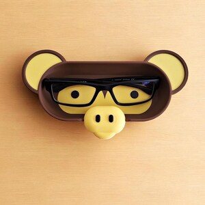 Puede incluir: Un original soporte para gafas con forma de cara de cerdo. El soporte es marr&oacute;n con detalles amarillos para las orejas, los ojos y la nariz. Unas gafas negras descansan dentro del soporte, dise&ntilde;ado para colocarse sobre una superficie plana.