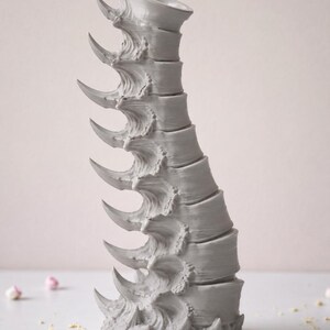 May include: A gray, sculptural vase with a spine-like design. The vase features a series of curved, pointed elements along its length, giving it a unique and textured appearance. The vase is on a white surface with scattered pink flower buds.
