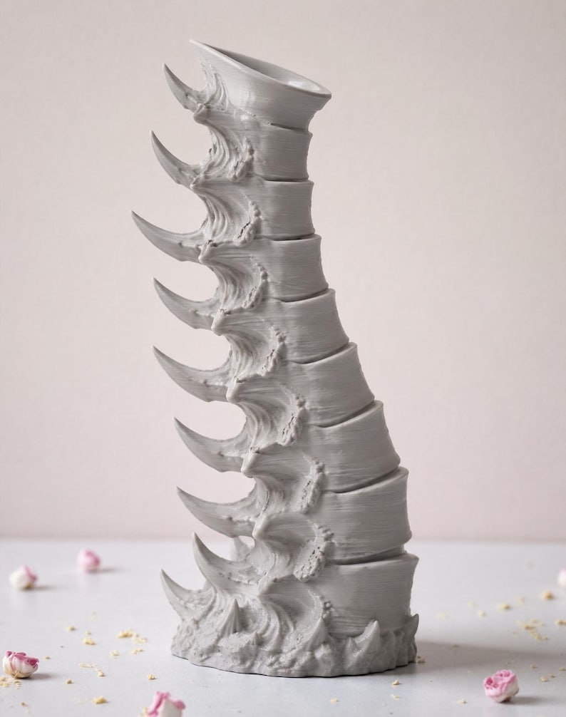 May include: A gray, sculptural vase with a spine-like design. The vase features a series of curved, pointed elements along its length, giving it a unique and textured appearance. The vase is on a white surface with scattered pink flower buds.