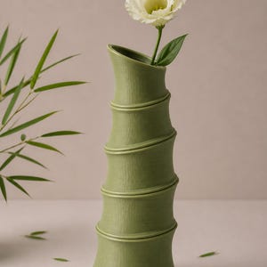 Puede incluir: Un jarr&oacute;n verde con forma de bamb&uacute; sostiene una flor blanca con un tallo y una hoja verdes. El jarr&oacute;n tiene una superficie texturizada y una abertura curva. El fondo es de un color suave y neutro, con hojas de bamb&uacute; esparcidas.