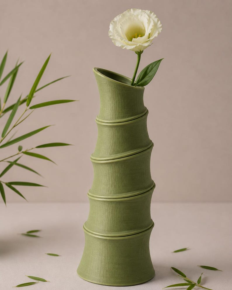 Puede incluir: Un jarr&oacute;n verde con forma de bamb&uacute; sostiene una flor blanca con un tallo y una hoja verdes. El jarr&oacute;n tiene una superficie texturizada y una abertura curva. El fondo es de un color suave y neutro, con hojas de bamb&uacute; esparcidas.